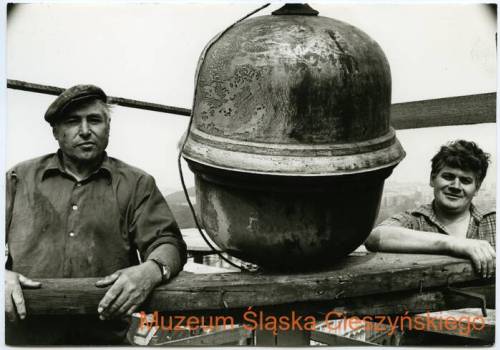 Czarno-białe zdjęcie przedstawiające dwóch mężczyzn pracujących przy remoncie wieży ratuszowej w 1984 r., a także montowaną wówczas banię. Zdjęcie autorstwa Tadeusza Popiela, z zasobów Muzeum Śląska Cieszyńskiego.fot.cieszyn.pl