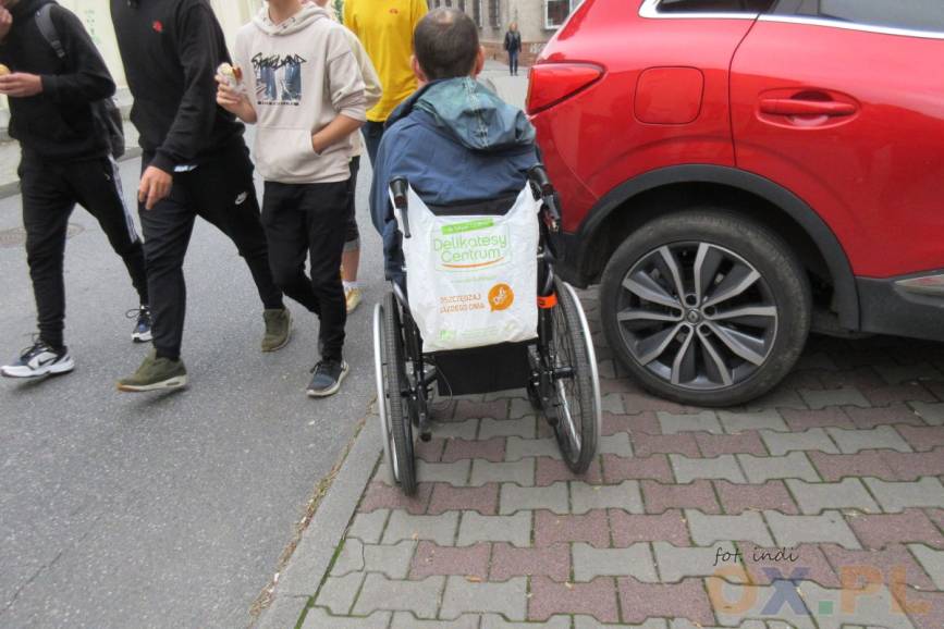 Osoba na wózku inwalidzkim próbująca przejechać obok auta które uniemożliwia osobie swobodnie przejechanie