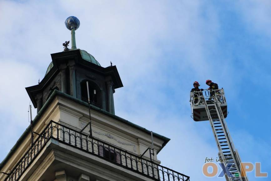 Zdjęcie przedstawia dwóch strażaków próbujących dostać się do iglicy na wieży Ratusza z drabiny.