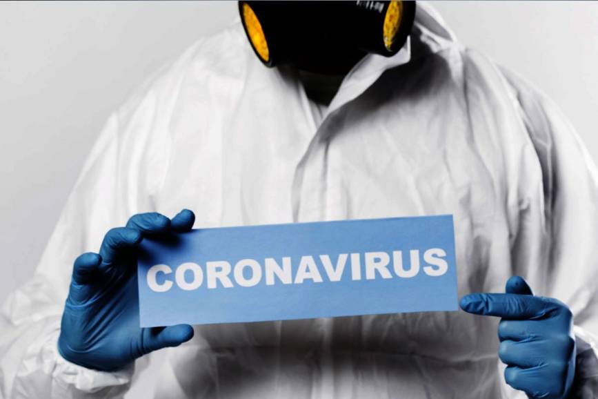 Człowiek ubrany w kombinezon ochronny, rękawiczki i maskę. W rękach trzyma tabliczkę z napisem CORONAVIRUS.