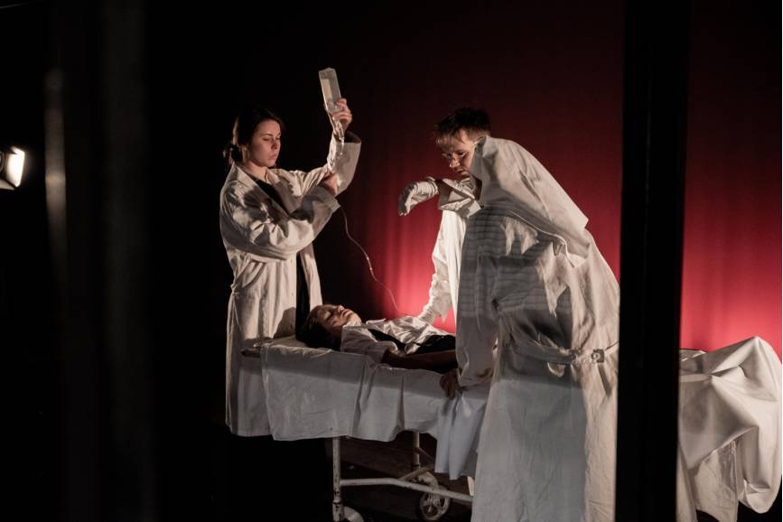 Zdjęcie przedstawia fragment spektaklu. 3 osoby, lekarz i pielęgniarki pochylają się nad chorym leżącym na łóżku szpitalnym