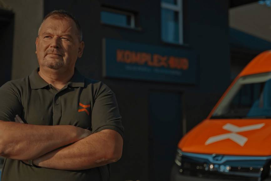Zdjęcie przedstawia z lewej strony na pierwszym planie mężczyznę w  koszulce z logo Komplex-Bud, na drugim planie z prawej strony fragment maski samochodu w barwach pomarańczowych z logo Komplex-Bud oraz pośrodku w tle logo firmy, szyld na budynku