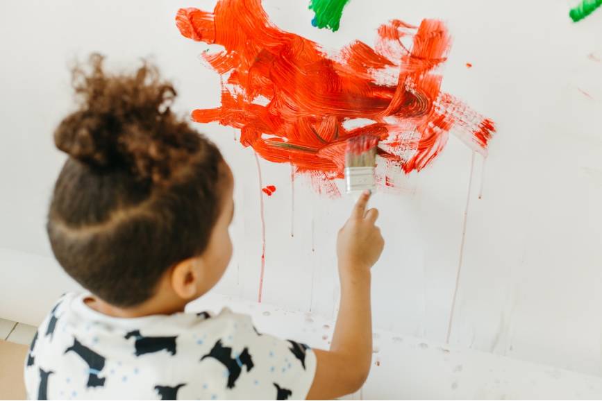Zdjęcie przedstawia dziecko stojące tyłem do sztalugi, na której maluje pędzlem