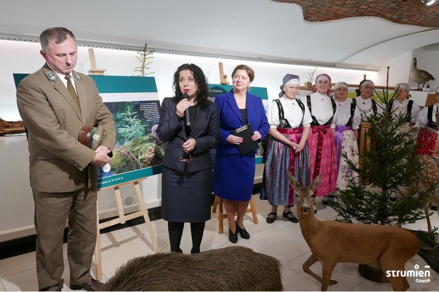 Na zdjęciu Burmistrzyni Strumienia Anna Grygierek otwiera wystawę, w ręku trzyma mikrofon, obok niej przedstawiciele nadleśnictwa kobiór oraz zespół folklorystyczny 