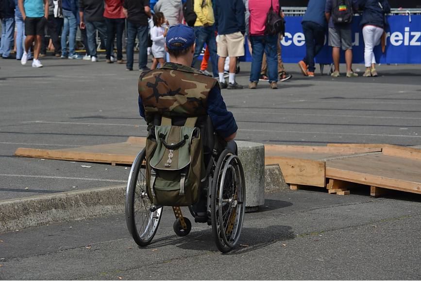 Osoba na wózku przed drewnianą przeszkodą na ulicy. Z tyłu wózka ma przyczepiony plecak. Czapka na głowie niebieska założona daszkiem do tyłu. Młoda niepełnosprawna osoba na zdjęciu